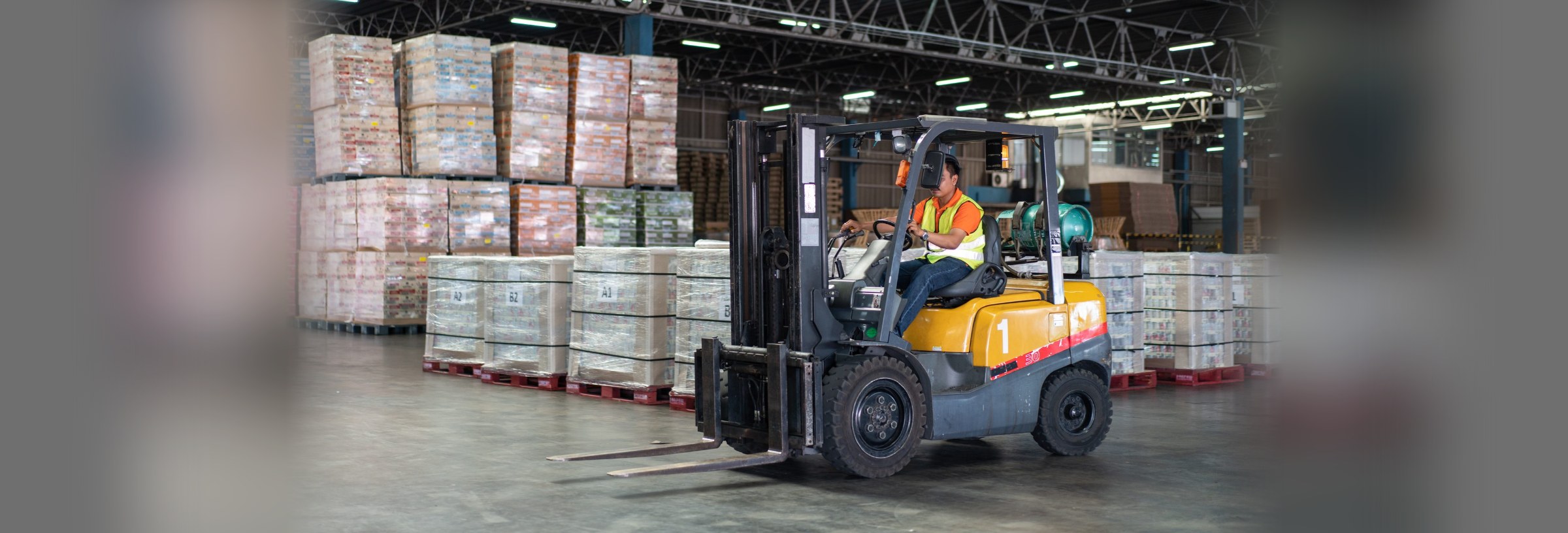 A forklift operator working in a large warehouse, driving past stacked pallets.