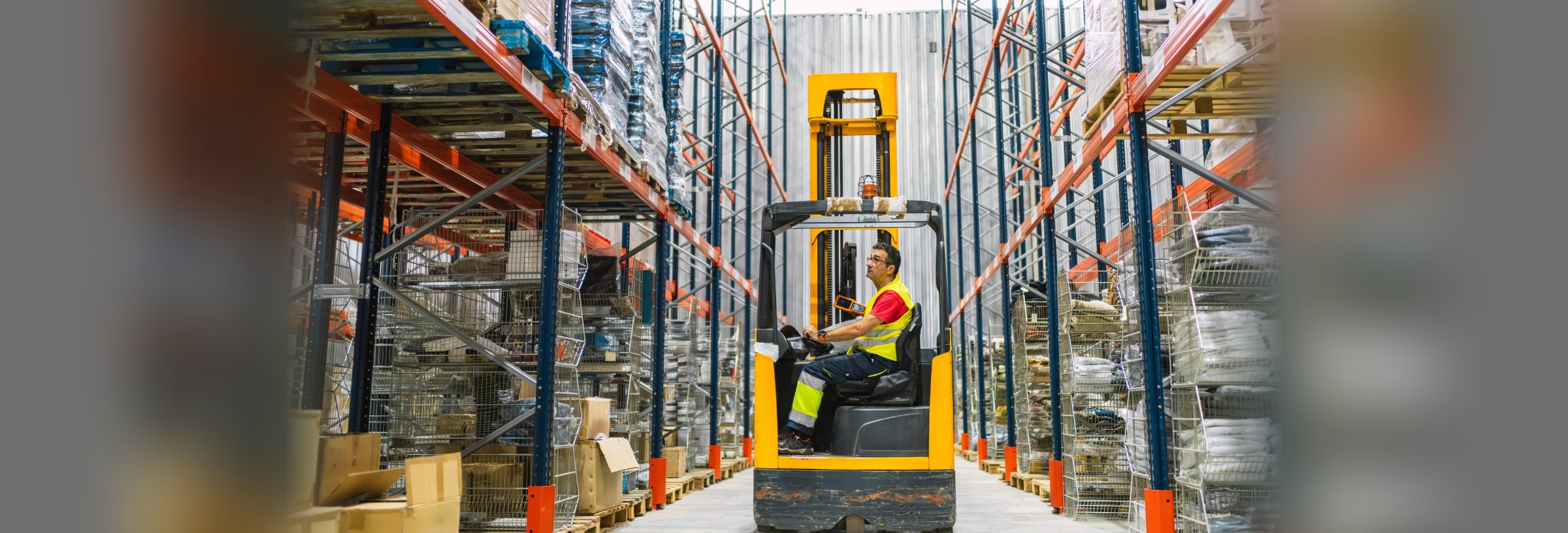 A forklift operator driving through a warehouse aisle, moving supplies past racked pallets.
