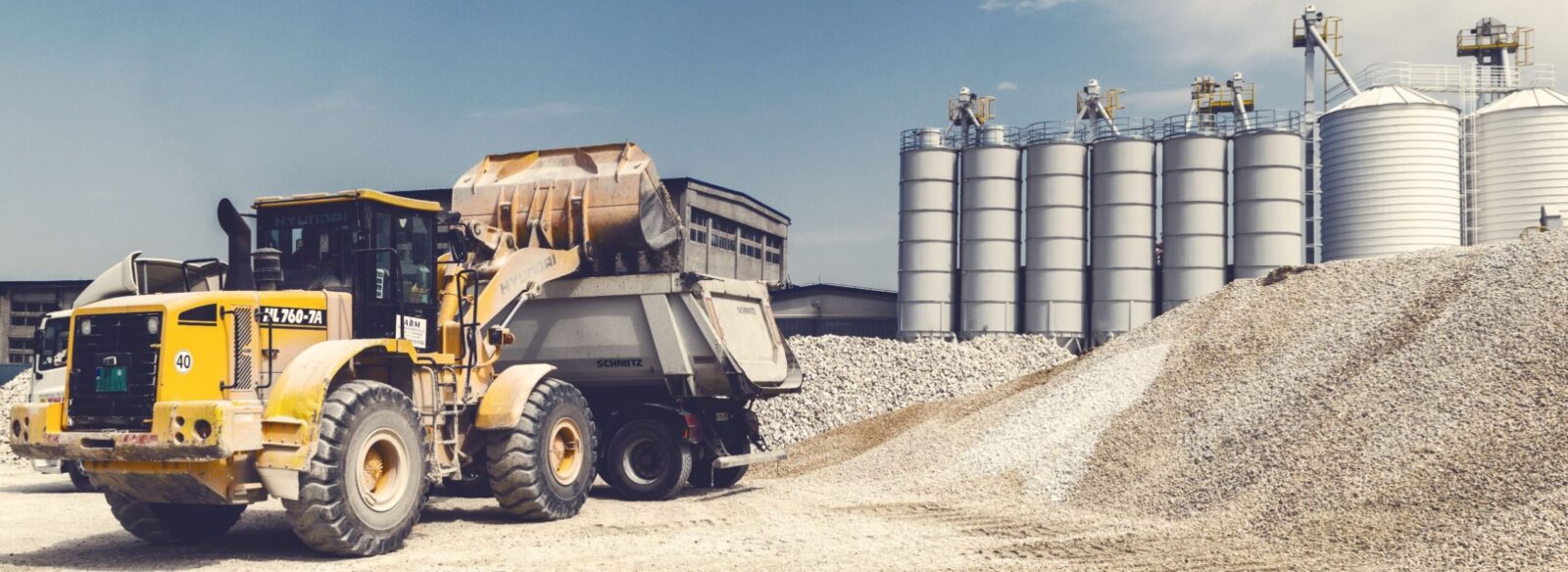 A wheel loader loading crushed aggregate into a tipper beside stockpiles, with storage silos behind.