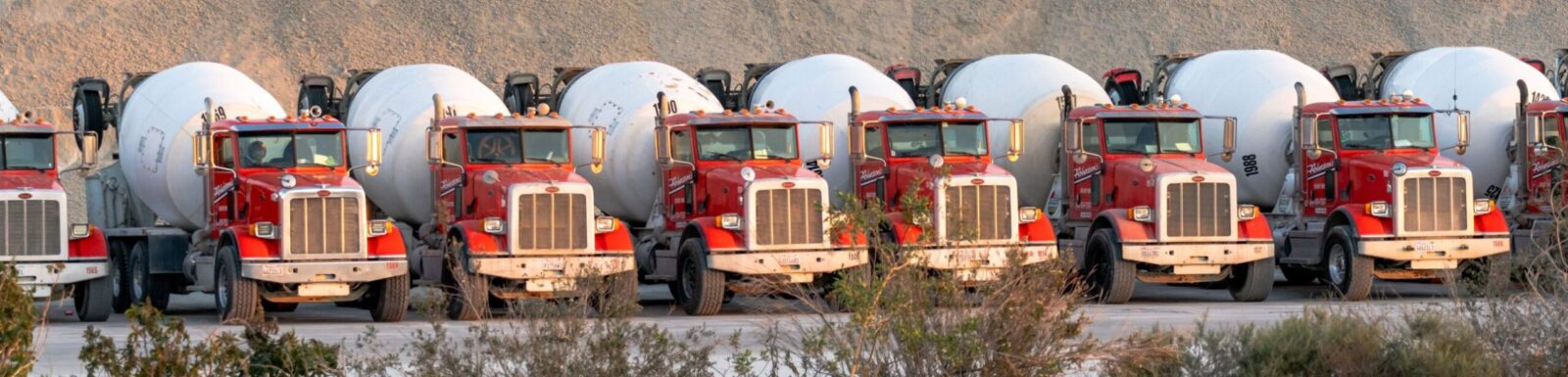 A line of ready-mix concrete mixer trucks parked at a plant yard, with an aggregate stockpile behind them.